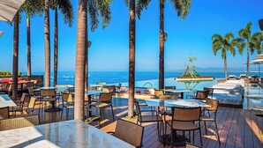 Uma área de refeições ao ar livre com mesas e cadeiras, cercada por palmeiras, com vista para uma piscina e o oceano.