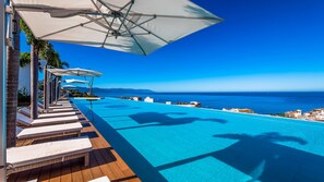 Un espace aménagé au bord de la piscine, avec des chaises longues et des parasols, offrant une vue sur une mer d’un bleu limpide et sur des montagnes lointaines.