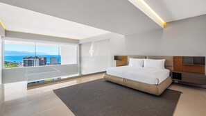 A modern bedroom with a large bed, a nightstand, a TV, and a view of the ocean.