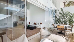 A modern interior with a glass partition separating a living area from a kitchen. There is a white sofa with brown cushions, a coffee table, and wicker chairs.