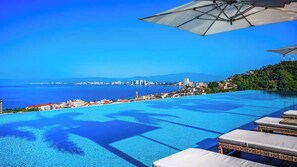 Una piscina infinita con vistas a una ciudad costera, sillas de descanso y sombrillas.