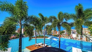 Une vue sur la piscine, avec des palmiers, des chaises longues et un ciel d’un bleu limpide.