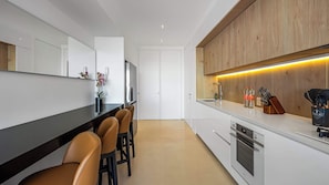 Eine moderne Küche mit schwarzem Arbeitsplatz, weißen Schränken und einer Wand in Holzoptik. Ein Barbereich bietet Platz für vier Stühle, und in der Arbeitsfläche befindet sich ein Spülbecken mit einem Messerblock.