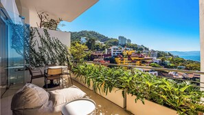 A balcony with a glass wall, a table with chairs, and a view of a coastal cityscape with buildings and greenery.