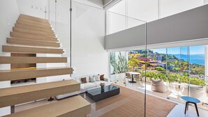 A modern living room with a glass staircase, a sofa, a coffee table, and a view of a coastal town.