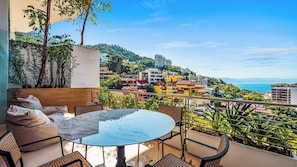 Ein Balkon mit Glastisch und Stühlen, der Blick auf eine Küstenstadt mit Gebäuden und Grünflächen bietet.