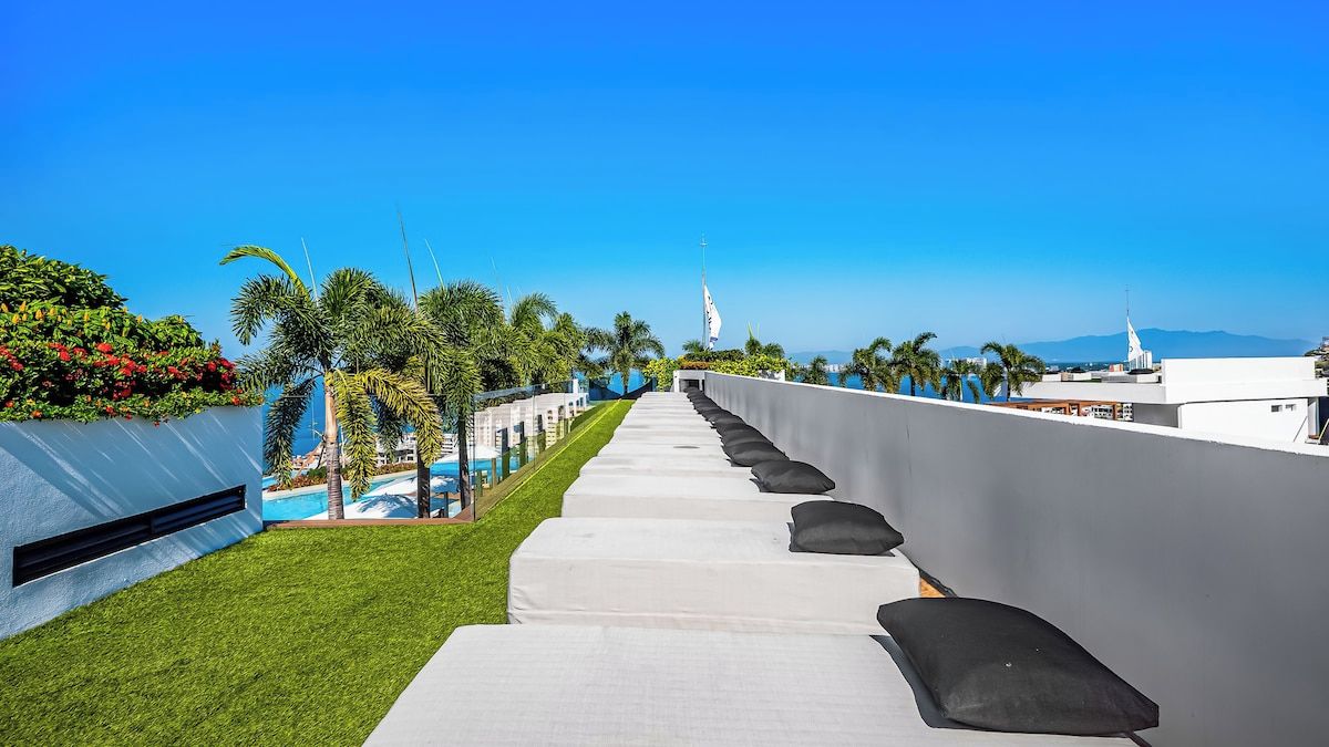 A rooftop terrace with a clear view of the ocean and mountains, featuring a long row of white beds with black pillows.