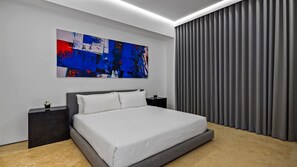 A modern bedroom with a large bed, a nightstand, a wall-mounted abstract painting, and a window with grey curtains. - AlilaHolidays - Luxury Getaway in SOHO PV - 105 (Puerto Vallarta)
