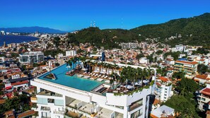 A modern hotel with a rooftop pool, surrounded by dense urban buildings and a clear view of the sea.