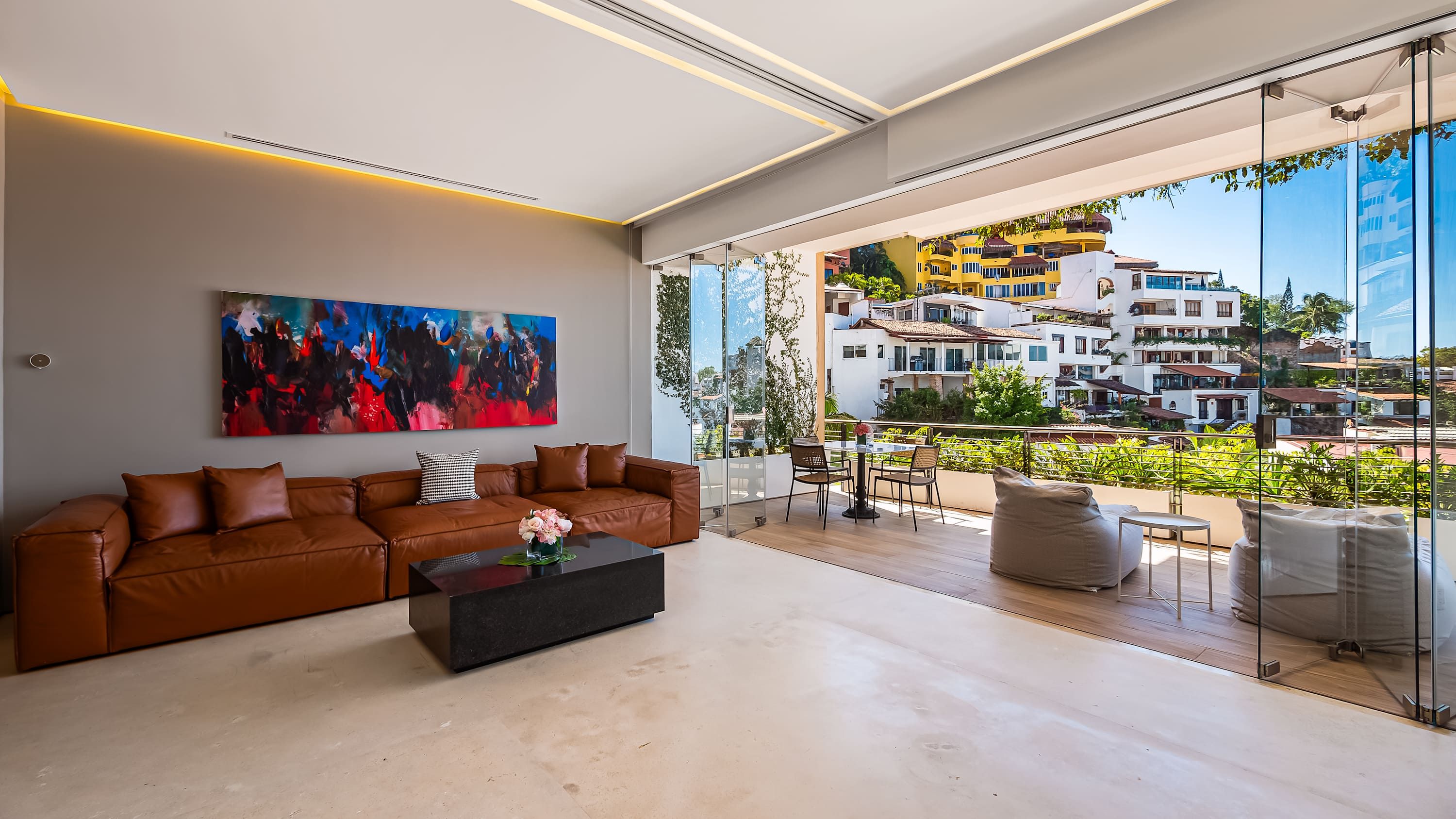 A modern living room with a large window, a brown leather sofa, a coffee table, and a colorful abstract painting.