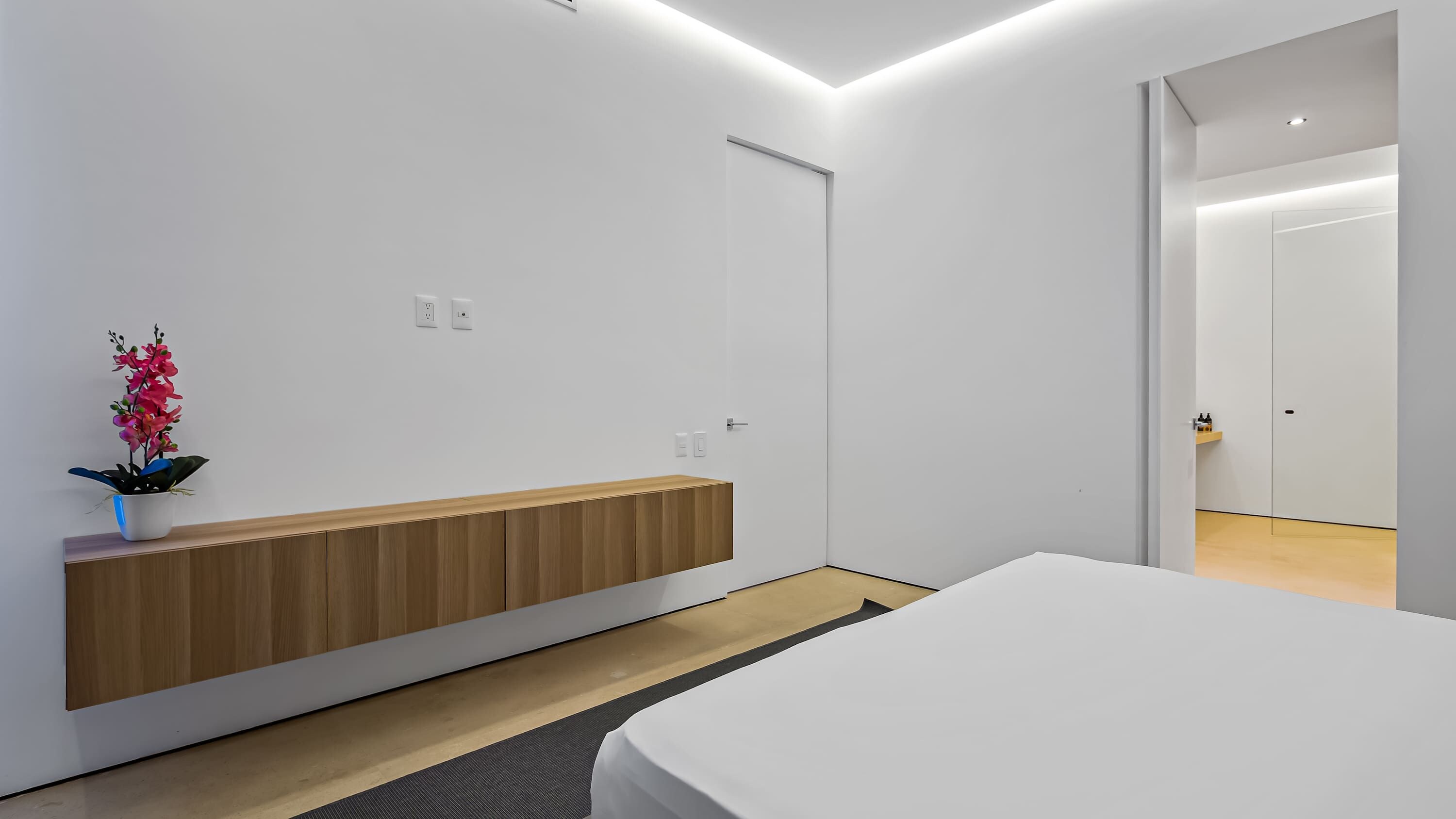 Moderni makuuhuone, jossa on puinen yöpöytä, valkoinen sänky ja ruukussa oleva kasvi.