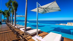 A poolside area with lounge chairs and umbrellas, overlooking a clear blue sea and distant buildings. - SoHo PV - 411 (Puerto Vallarta)