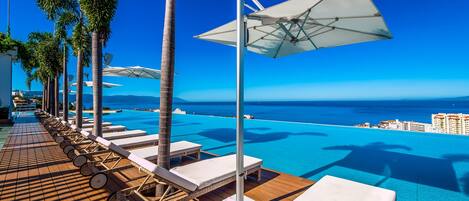 A poolside area with lounge chairs and umbrellas, overlooking a clear blue sea and distant buildings.