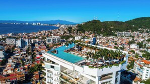 A coastal cityscape with a prominent rooftop pool area, modern buildings, and a view of the sea and distant mountains.