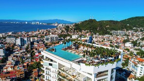 A coastal cityscape with a prominent rooftop pool area, modern buildings, and a view of the sea and distant mountains.