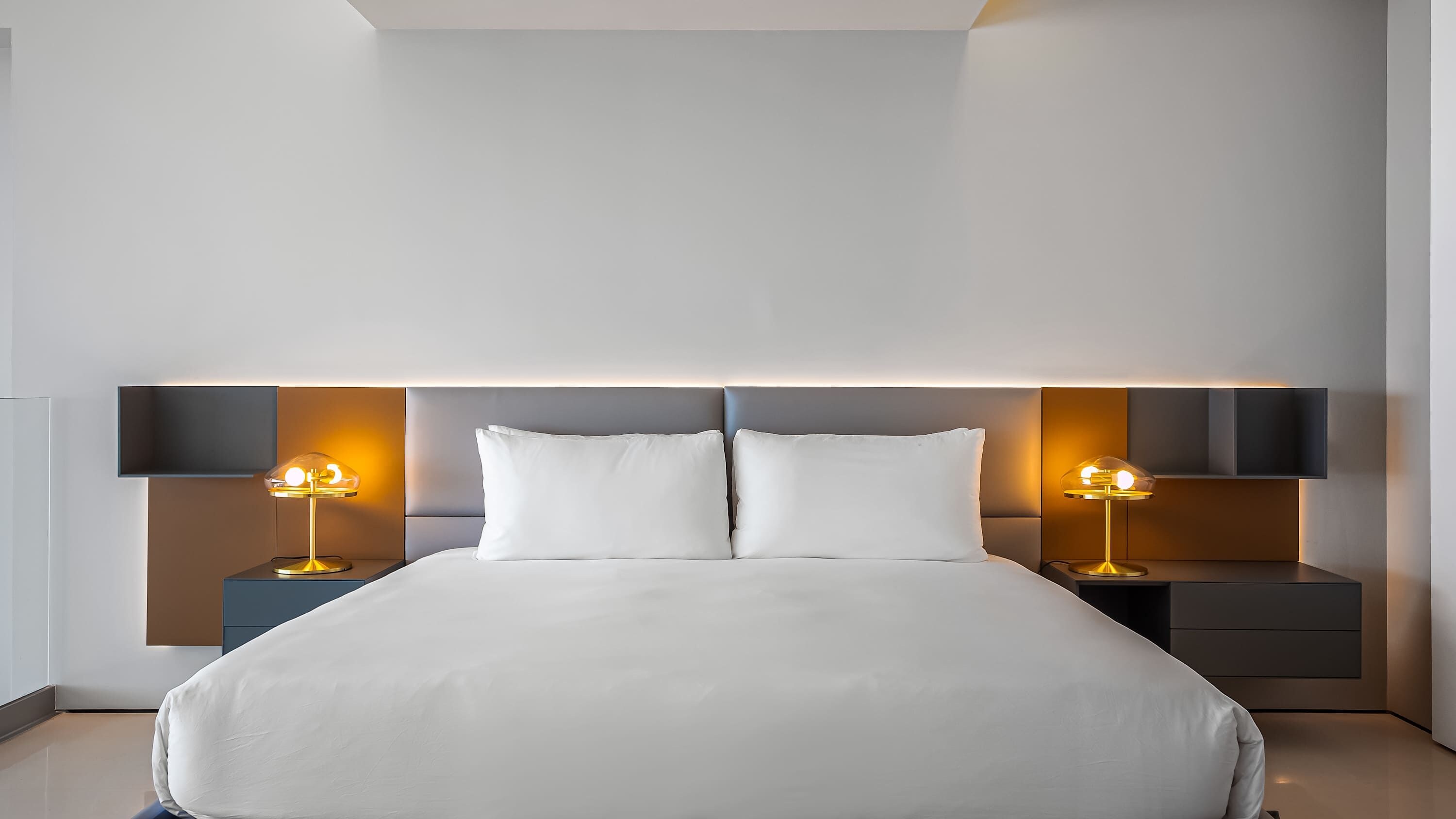A modern hotel room with a large bed, two bedside tables with lamps, and a headboard with built-in lighting.
