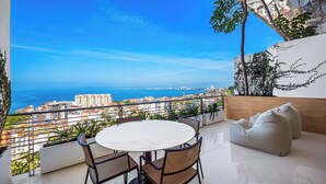 A balcony with a round table and chairs, overlooking a cityscape and the sea.