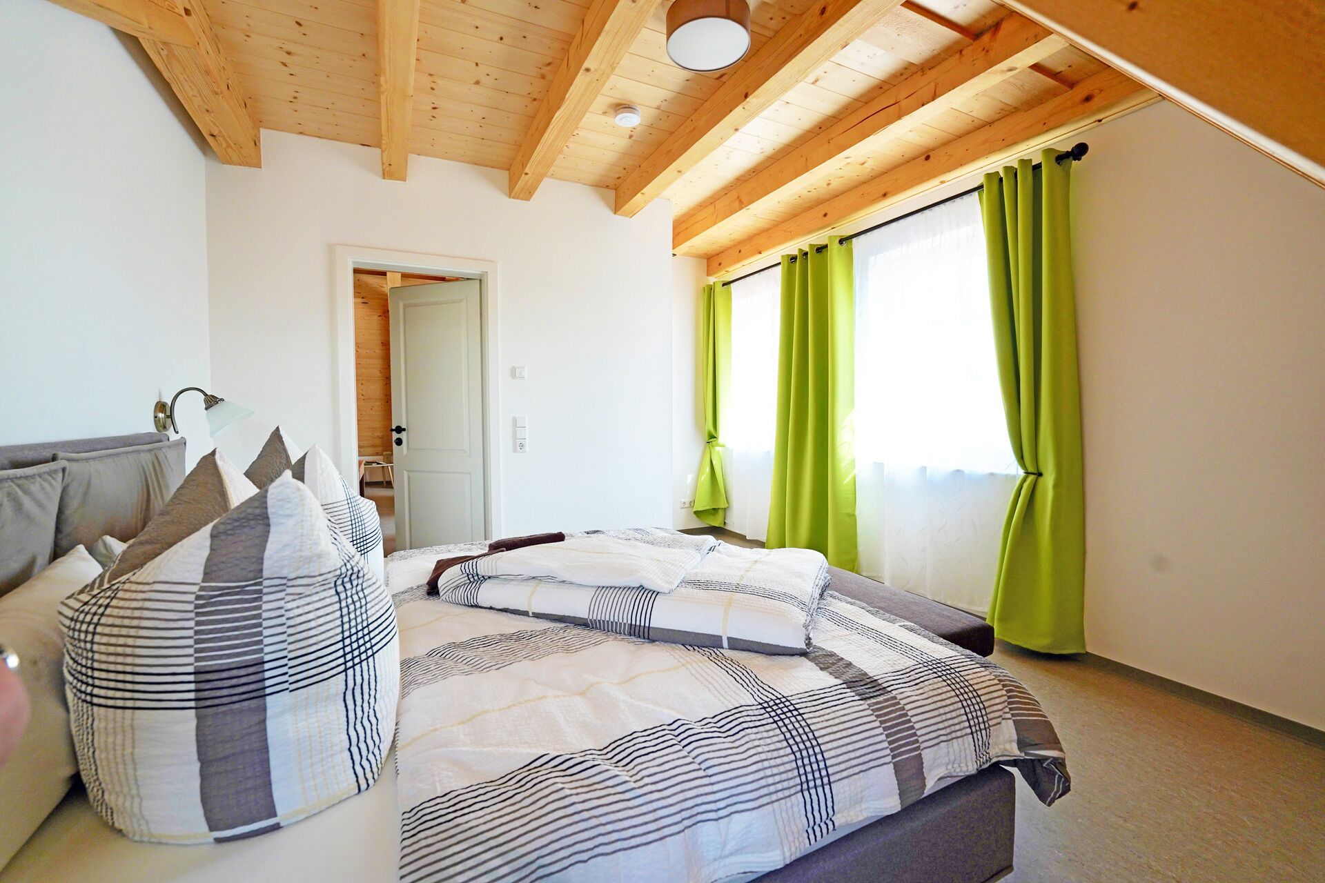 A bedroom with a bed, pillows, a lamp, a window with curtains, and a wooden ceiling.