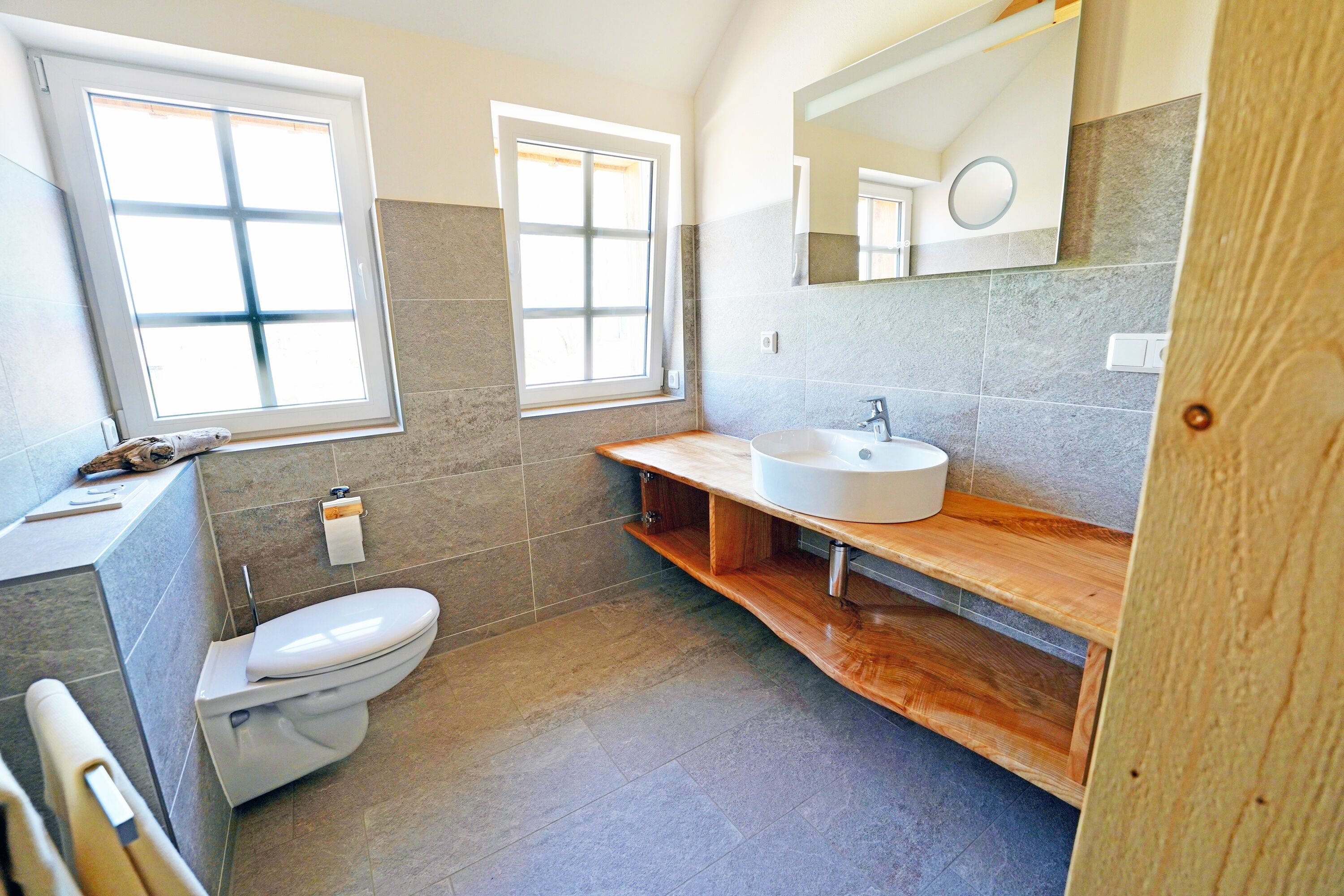 Un baño con inodoro, lavamanos, espejo y dos ventanas.