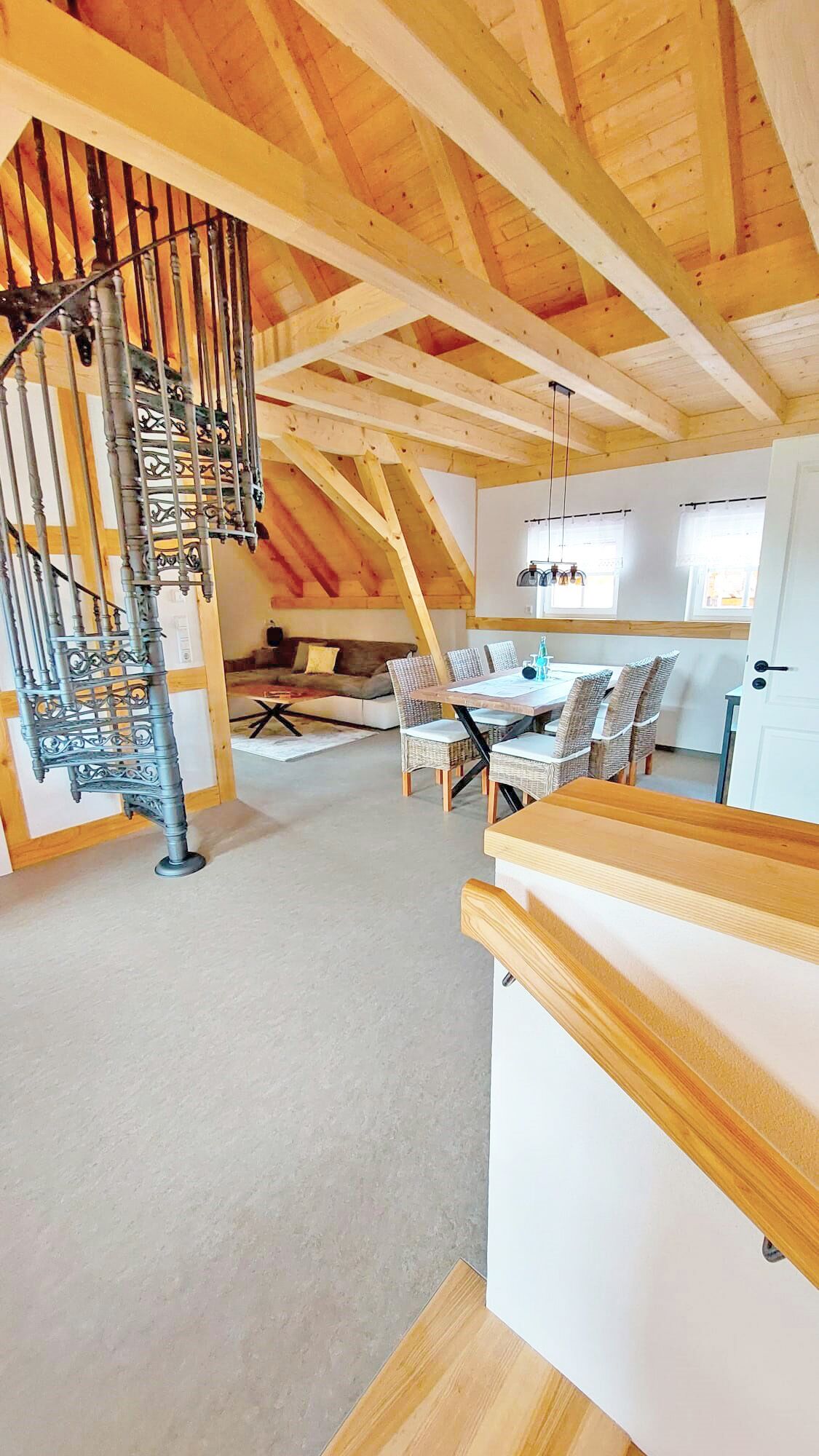 Un espacio tipo loft con techo de madera, una escalera de caracol, una zona de comedor con sillas de mimbre y un sofá.