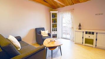 Ein Wohnzimmer mit Sofa, Sessel und Couchtisch, einem Bücherregal und einer Kommode mit kleinem Fenster.