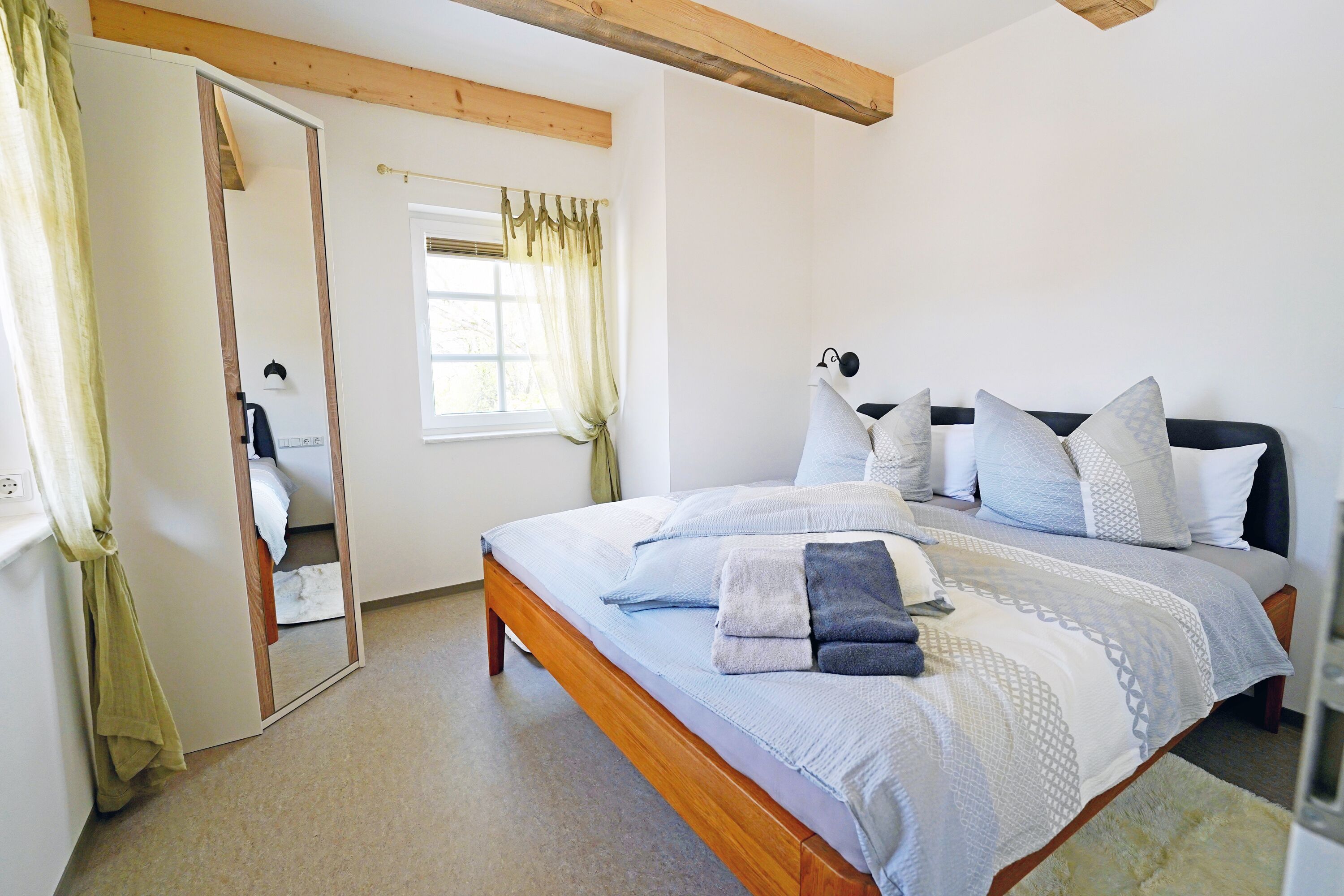 A bedroom with a wooden bed frame, a large mirror, a window with curtains, and a bedside table.