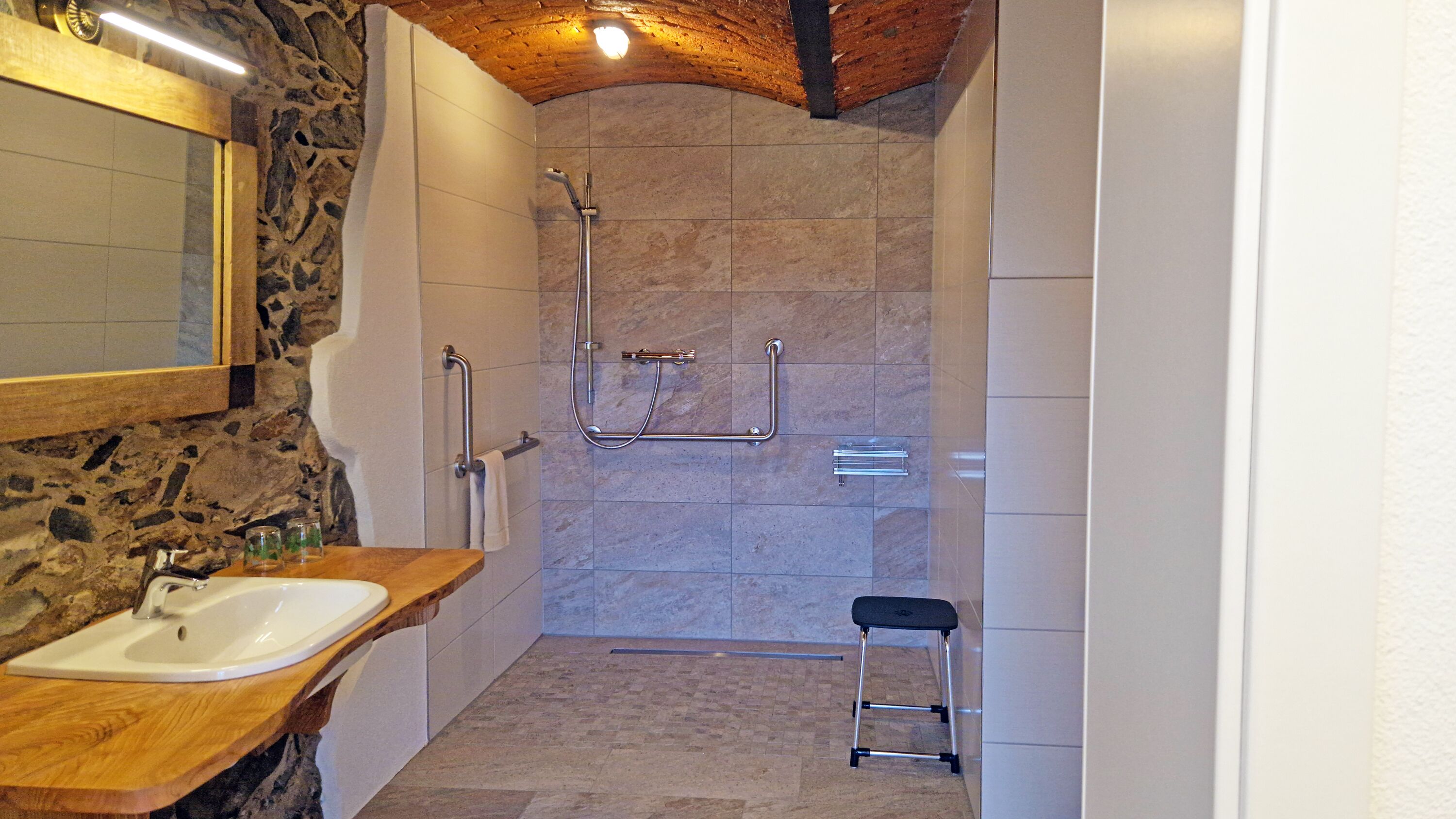 A bathroom with a stone wall, a wooden vanity, a shower with grab bars, and a small stool.