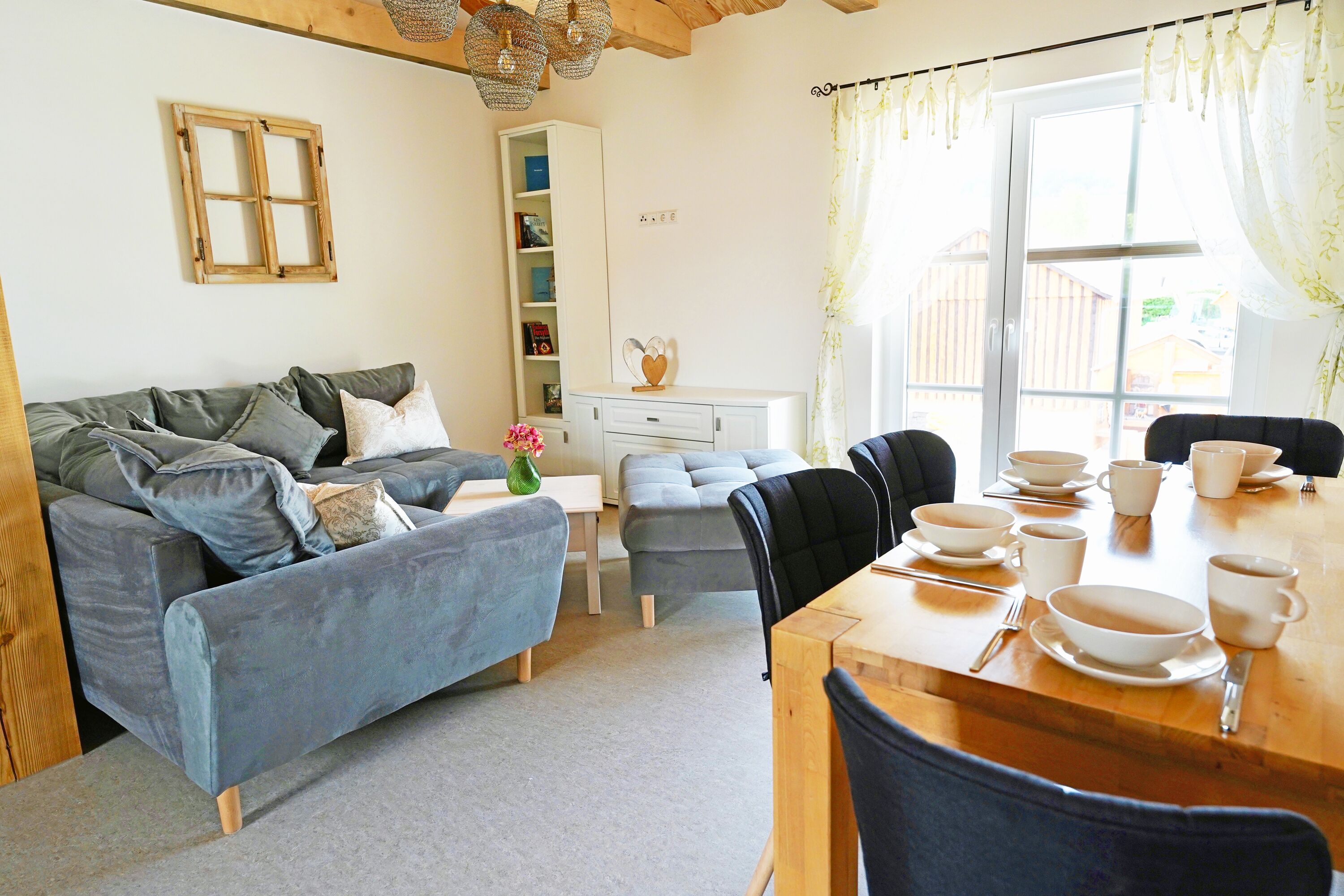 A cozy living room with a sofa, a dining table set with bowls and cups, a bookshelf, and a window with curtains.