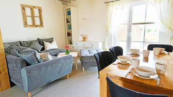 A cozy living room with a sofa, a dining table set with bowls and cups, a bookshelf, and a window with curtains.
