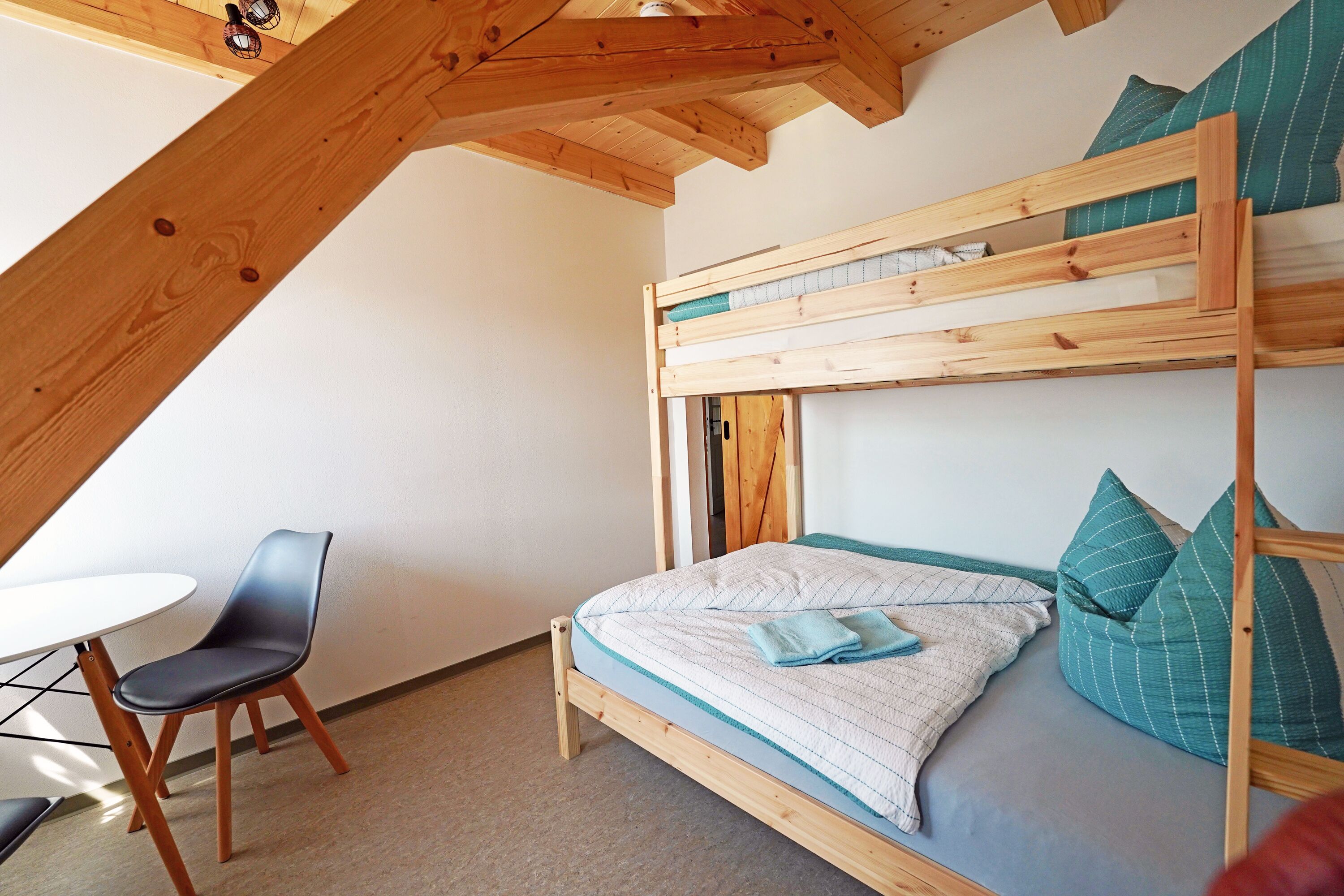 Letto a castello con struttura in legno e un letto singolo sottostante. C'è un tavolino con una sedia e un cuscino sul letto.