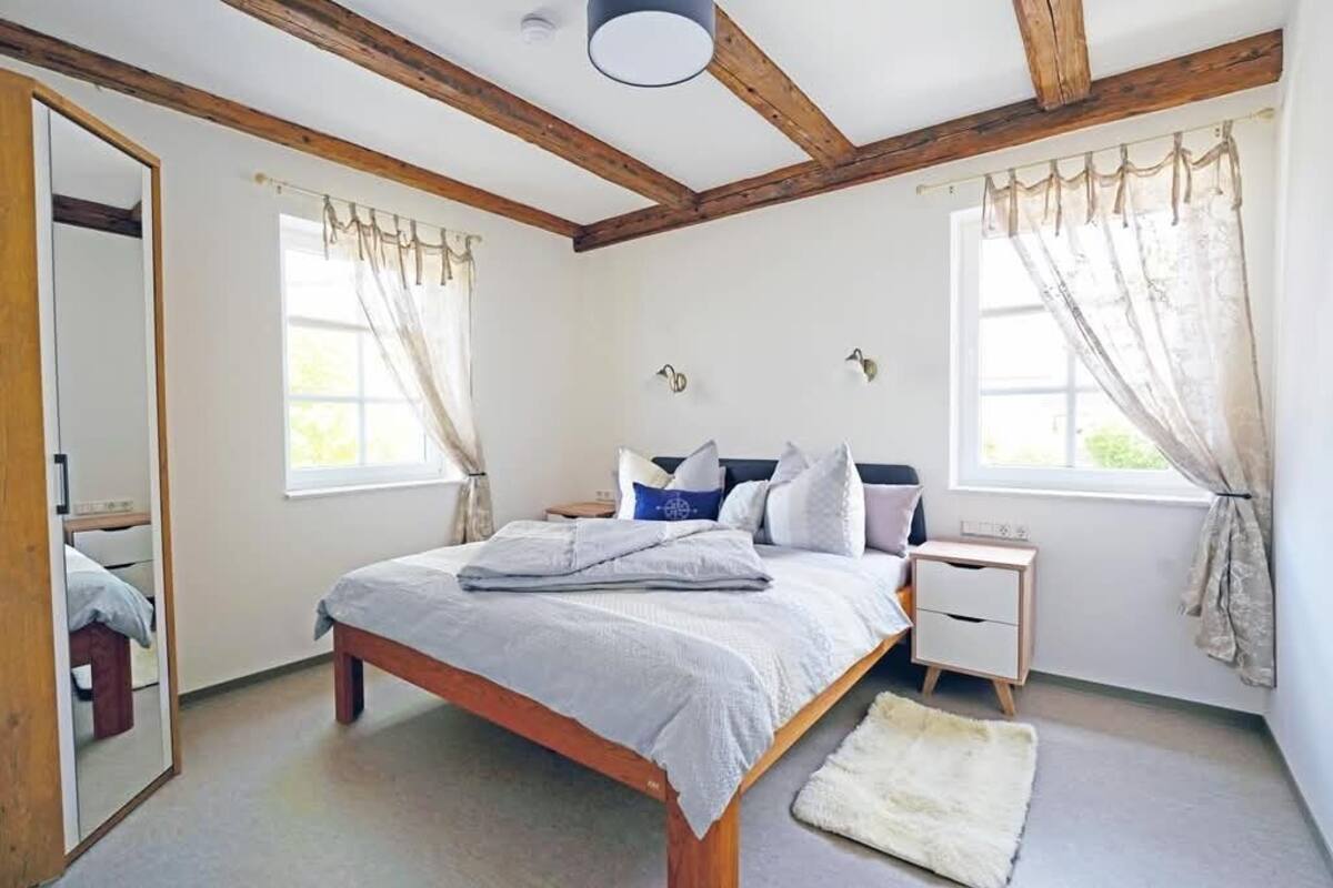 Un dormitorio con cabecero de madera, una mesita de noche, un espejo y una ventana con cortinas.