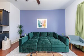 Una sala de estar con un sofá seccional verde, una mesita auxiliar, una planta en maceta y una obra de arte abstracta colgada en la pared.