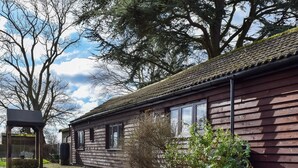 A wooden house with a tiled roof, a paved driveway, and a garden with various plants. - Brooklands Farm Cottage (Biggleswade)