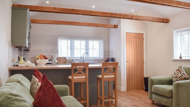 A modern kitchen with wooden beams, a dining area with green sofas, and a view of the outside. - Brooklands Farm Cottage (Biggleswade)