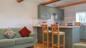 A cozy kitchen with a green sofa, two wooden bar stools, and a window with blinds. - Brooklands Farm Cottage (Biggleswade)