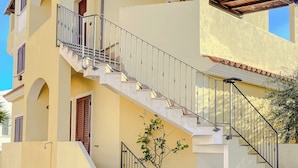 A two-story building with a stone-paved courtyard, a staircase with metal railings, and potted plants. - Porto Apartments - La Caletta (Siniscola)