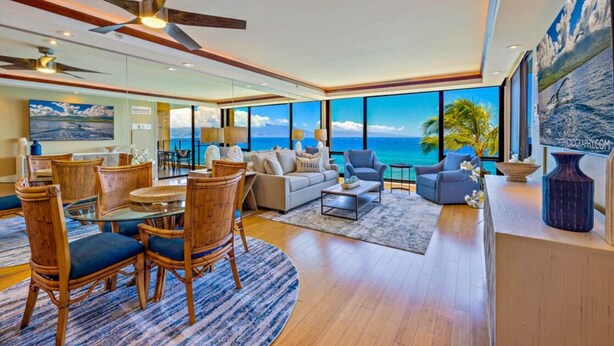 A spacious living room with a large window overlooking the ocean, a dining area with wooden furniture, and a modern ceiling fan.