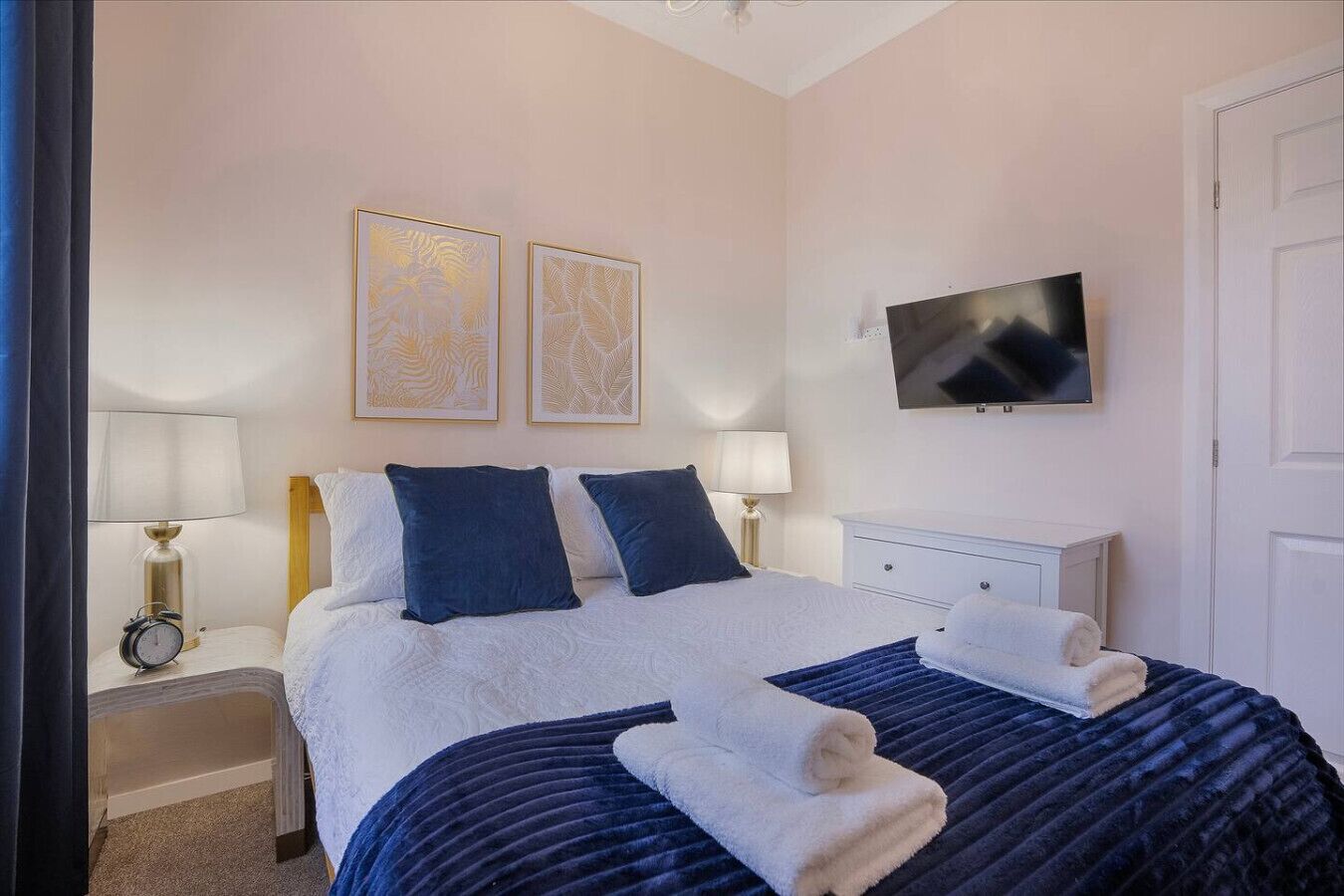 A neatly arranged bedroom with a bed, two bedside lamps, a flat-screen TV, and two framed artworks on the wall.