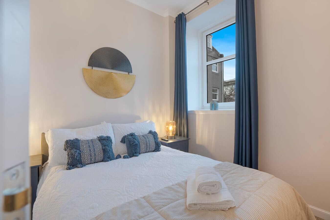 Cama bem arrumada com lençóis brancos e almofadas estampadas, luminária de cabeceira e janela com cortinas.