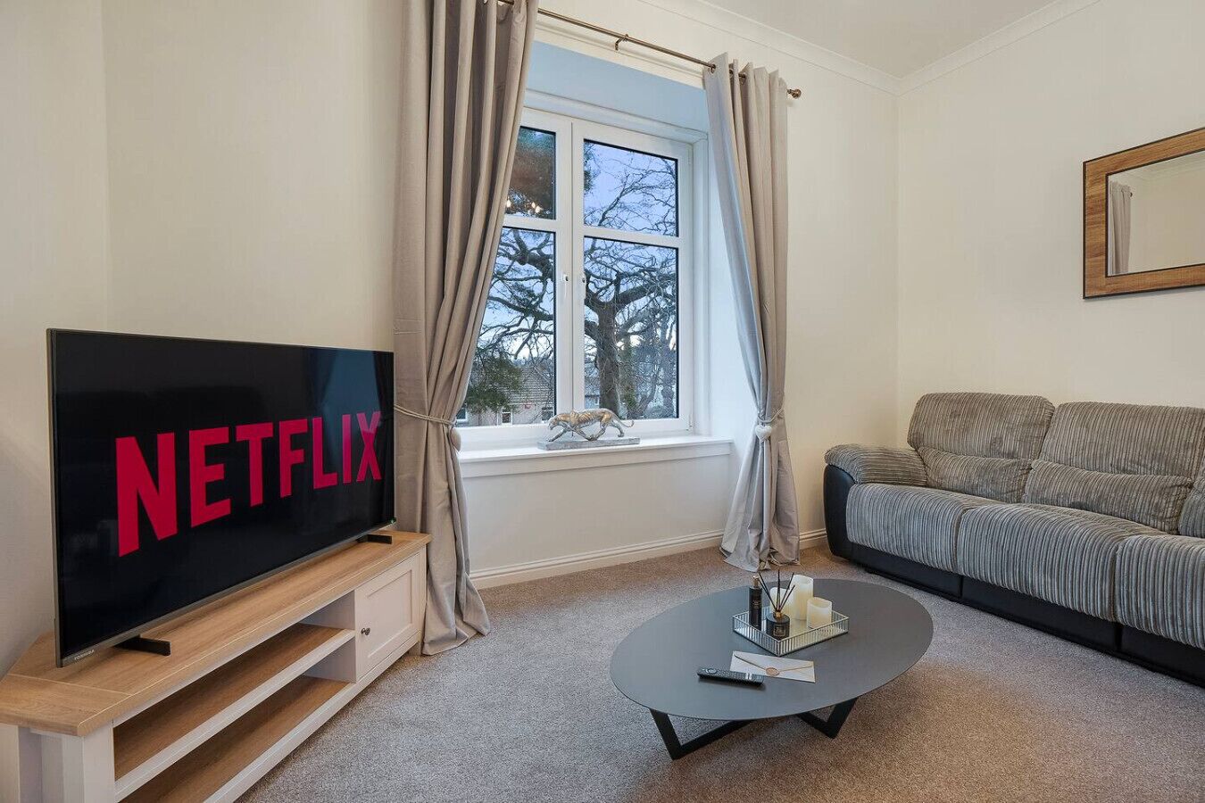 Ein Wohnzimmer mit einem großen Flachbildfernseher, der 'NETFLIX' zeigt, einer Couch, einem Couchtisch und einem Fenster mit Vorhängen.