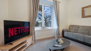 A living room with a large flat-screen TV displaying 'NETFLIX', a sofa, a coffee table, and a window with curtains. - Bethany House | Grampian Lettings (Aberdeen)