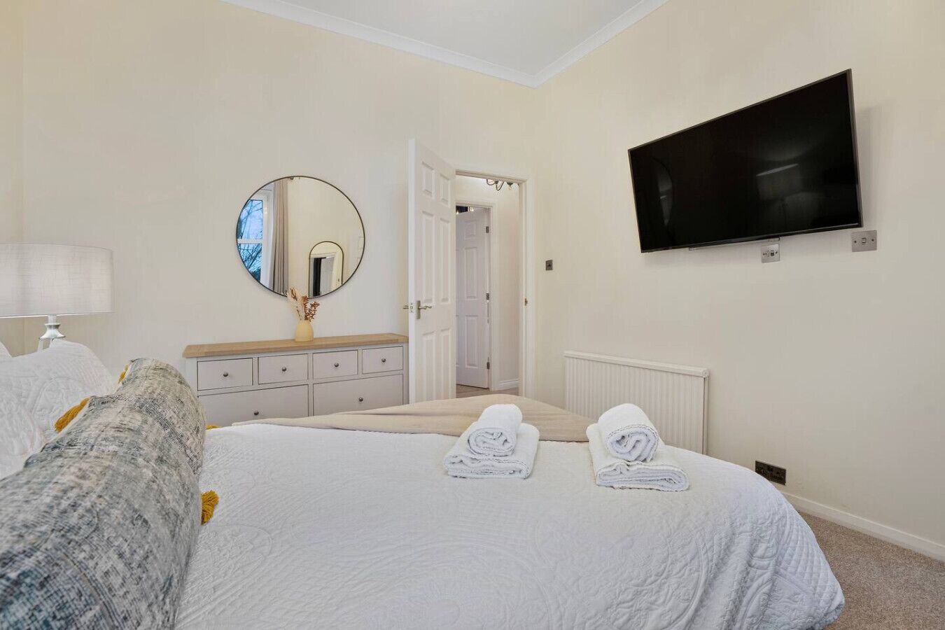 A hotel room with a bed, a bedside table with a lamp, a dresser, a mirror, a television, and a door leading to another room.
