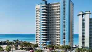 A high-rise hotel with multiple balconies, located near a beach with palm trees and a clear blue sky.