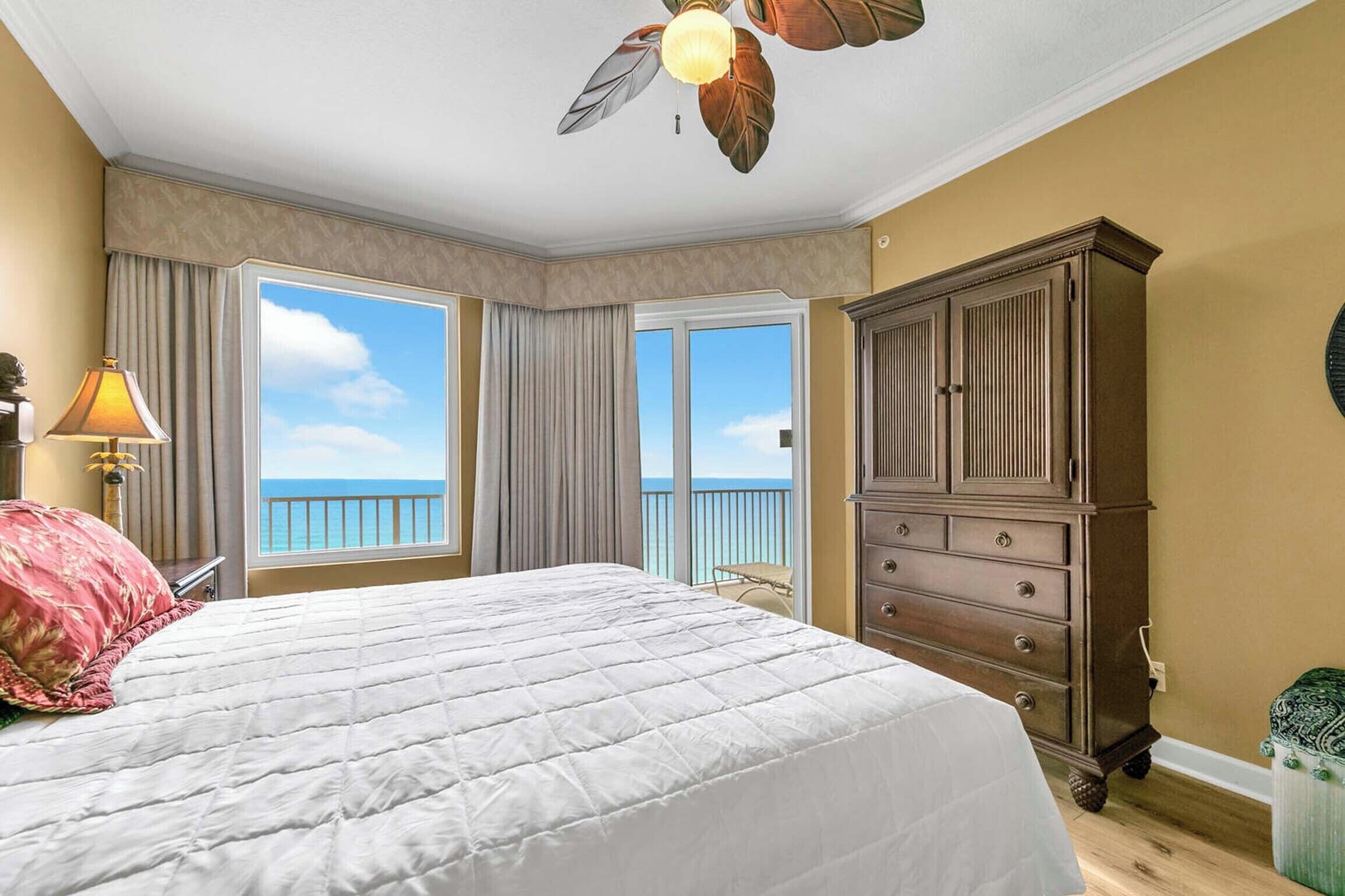 Ein Schlafzimmer mit einem großen Bett, einer Holzkommode, einem Deckenventilator und Blick auf den Ozean durch die Fenster.