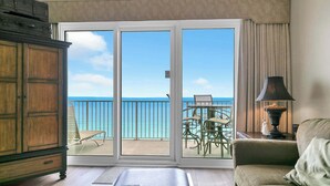 A room with a large sliding glass door leading to a balcony with a view of the ocean. There is a wooden cabinet, a sofa, and a table with a lamp.