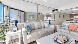A modern hotel room with a large window, a sofa, a coffee table, and a kitchen area visible through a glass partition. - ⛱️Stunning Beach Front Views-11 Pools-Leeward 403 at Edgewater⛱️ (Panama City)
