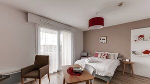 A modern hotel room with a bed, a desk, a chair, a red lamp, and a window with sheer curtains. - Zenao Appart'hôtels Challans (Challans)