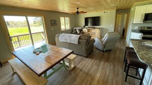 A spacious living room with a large sofa, a dining table, and a kitchen area with a TV and cabinets. - Rifle River Lodge, Rifle River Lodge LLC (Rose City)