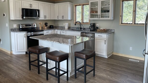 A modern kitchen with white cabinets, a granite countertop, and a central island with bar stools. - Rifle River Lodge, Rifle River Lodge LLC (Rose City)