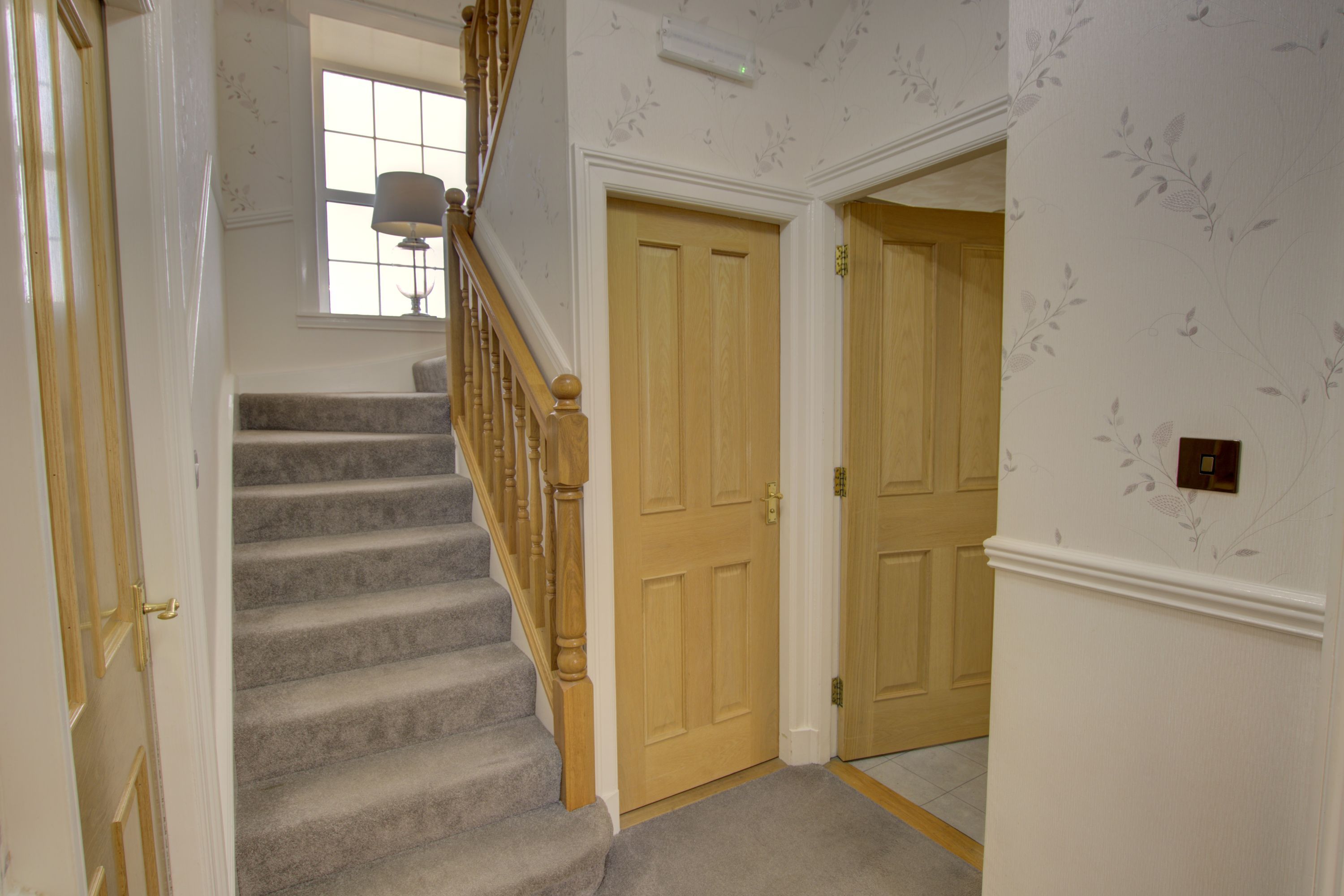 A carpeted staircase with wooden balusters, a light switch, and a door with a floral wallpaper pattern.